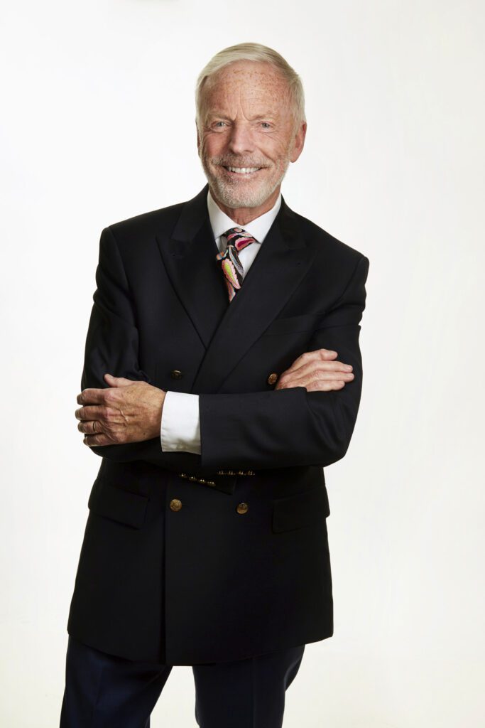 Business headshot of a smiling older man with white hair in a navy double-breasted suit with a patterned tie, arms crossed, photographed in Kitchener