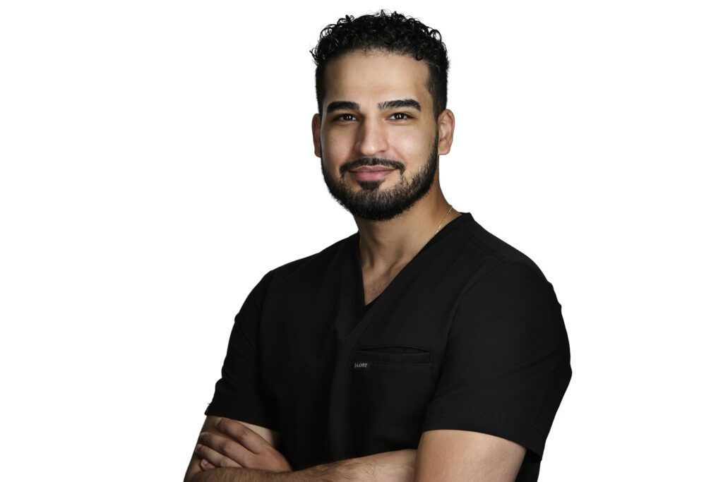 Dentist headshot of a man with a beard in black medical scrubs, arms crossed, taken on a white background