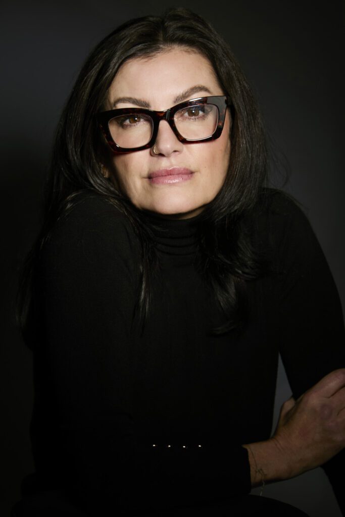 Model headshot of a woman with long dark hair and tortoiseshell glasses in a black turtleneck, dramatic dark backdrop, photographed in Kitchener