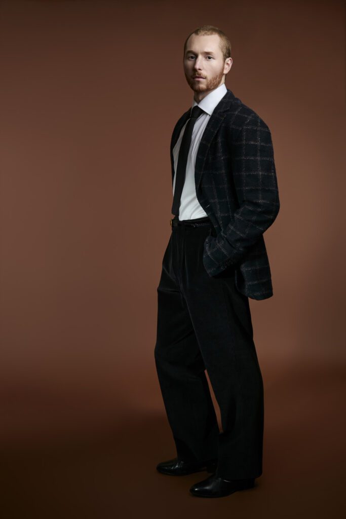 Waterloo model headshot: man in a checked blazer and dark trousers against a brown backdrop.